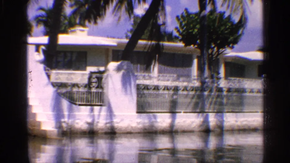 1959 California frame of a white waterside home with palm trees, exterior railing, low wall, and still water in front.