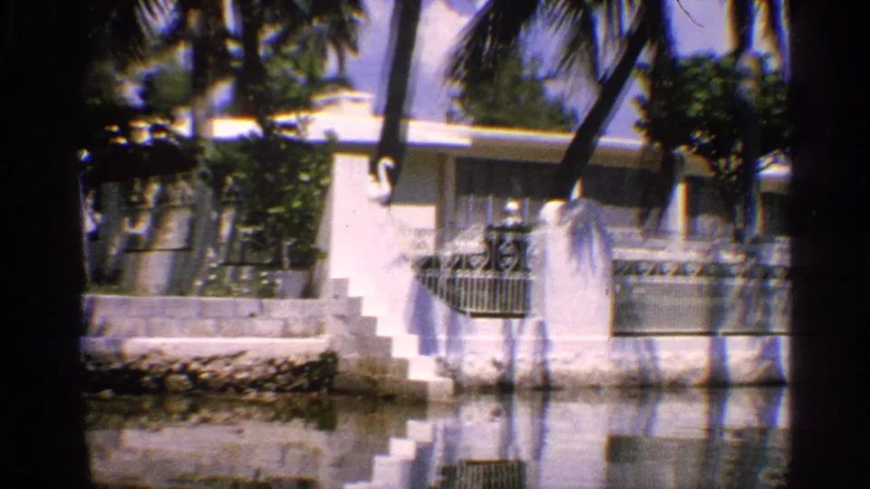 1959 California home-movie view of a waterfront house with exterior stairs, railings, palm trunks, and reflections in the water.