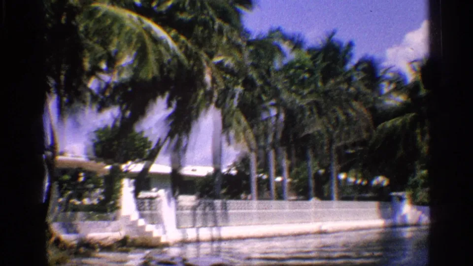1959 California frame showing palms over a white waterfront property, exterior steps, low wall, and rippled river water.