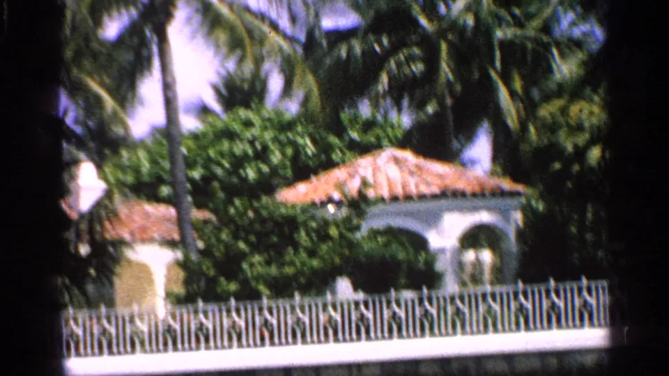 1959 Florida exterior view of a white stucco house with an orange terra cotta roof, palms, and white fencing.