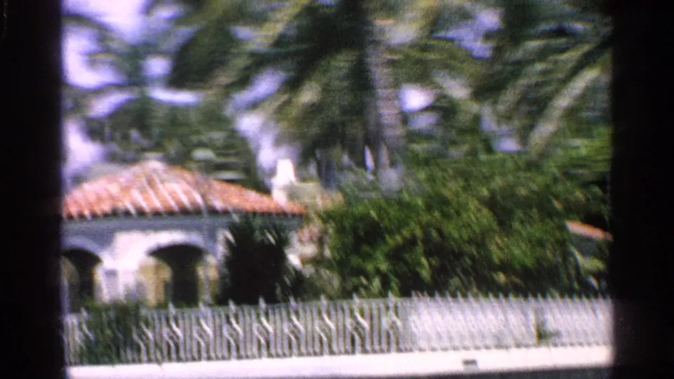 1959 Florida home-movie frame with motion blur over a white house, terra cotta roof, fence, palms, and shrubs.