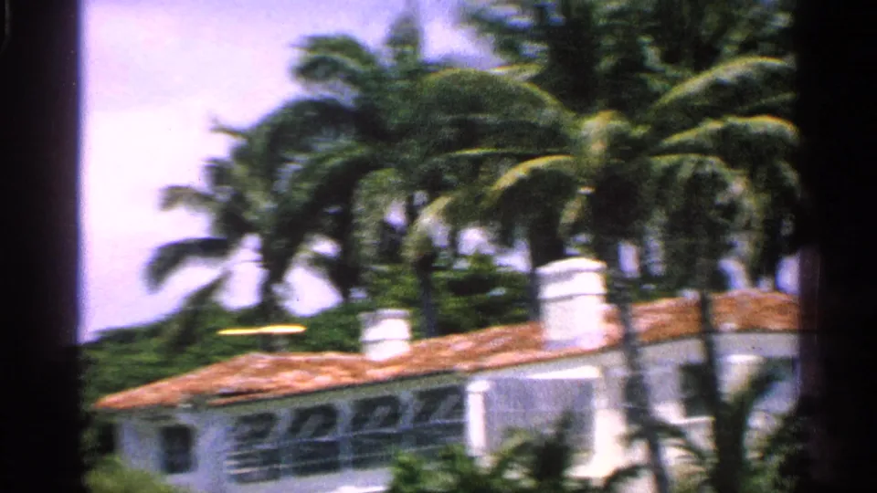 1959 Florida 8mm frame showing a white stucco house and terracotta roof seen through palm trees from a moving viewpoint.
