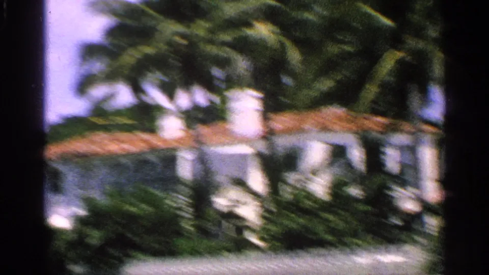 1959 Florida home-movie frame with motion blur across a white stucco building, terracotta roof, palms, shrubs, and a faint water glimpse.
