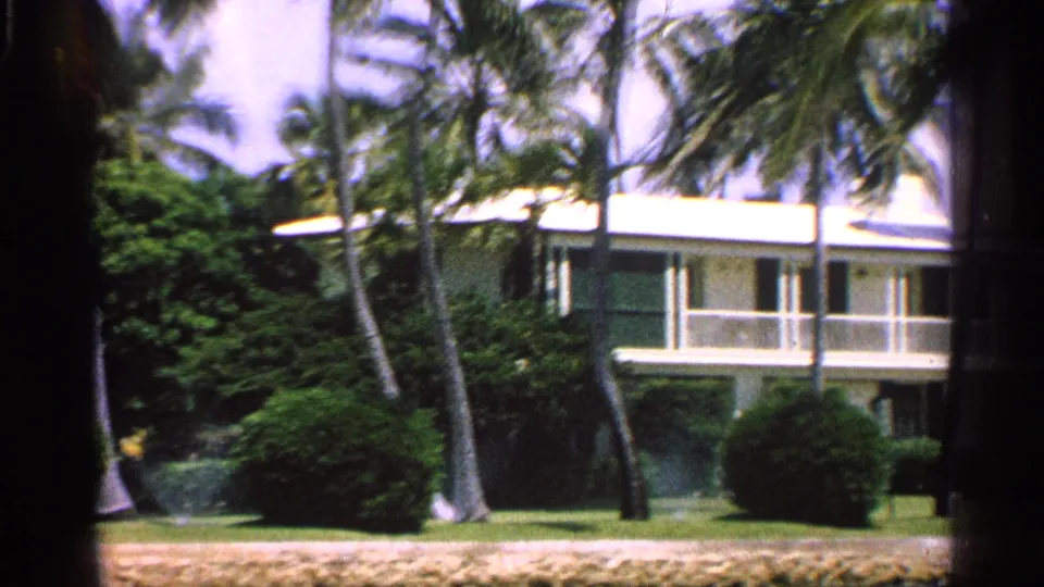 1959 Florida 8mm frame showing a two-story residence with a white balcony, palms, hedges, and sunlit lawn.