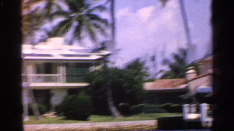 1959 Florida drive-by frame with motion blur across a balcony residence, palm trees, roof tiles, and a partial vehicle edge.