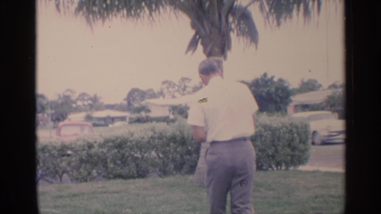 In 1959 Florida home-movie footage, a man in a white shirt stands with his back to camera on a grassy lawn near hedges and a palm tree.