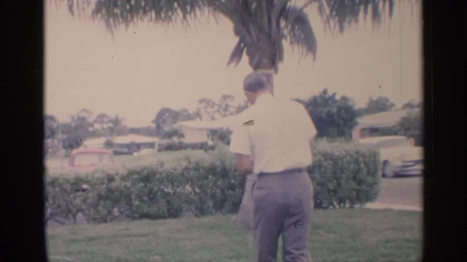 In 1959 Florida home-movie footage, a man in a white shirt stands with his back to camera on a grassy lawn near hedges and a palm tree.