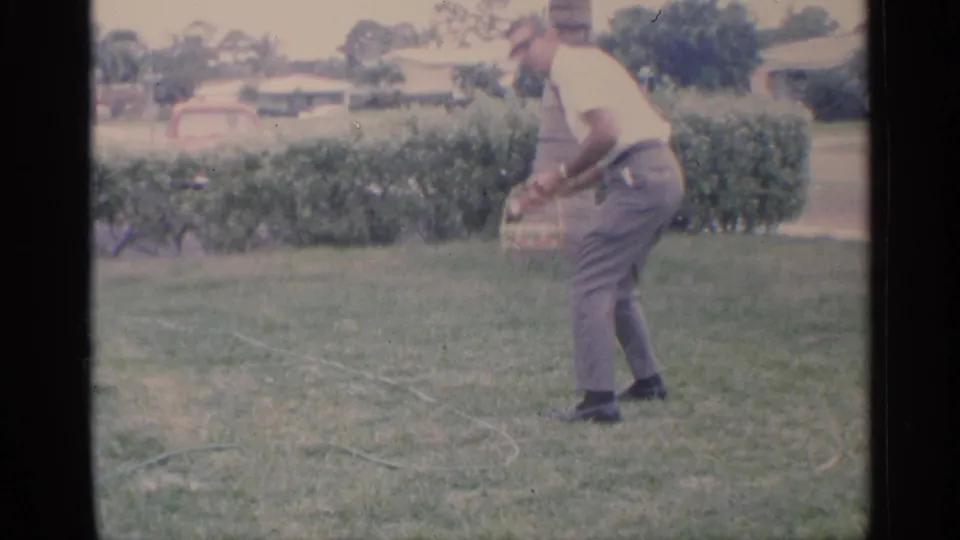 The man bends toward the grass with a small basket in hand during a Florida Easter egg hunt, with hedges and houses in the background.