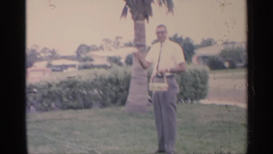 A man in a white shirt and tie stands beside a palm tree holding a small basket in a 1959 Florida yard scene.