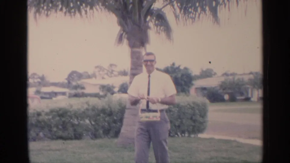 The man faces the camera in a Florida suburban yard, holding his Easter basket against a backdrop of hedges, palm tree, and street.
