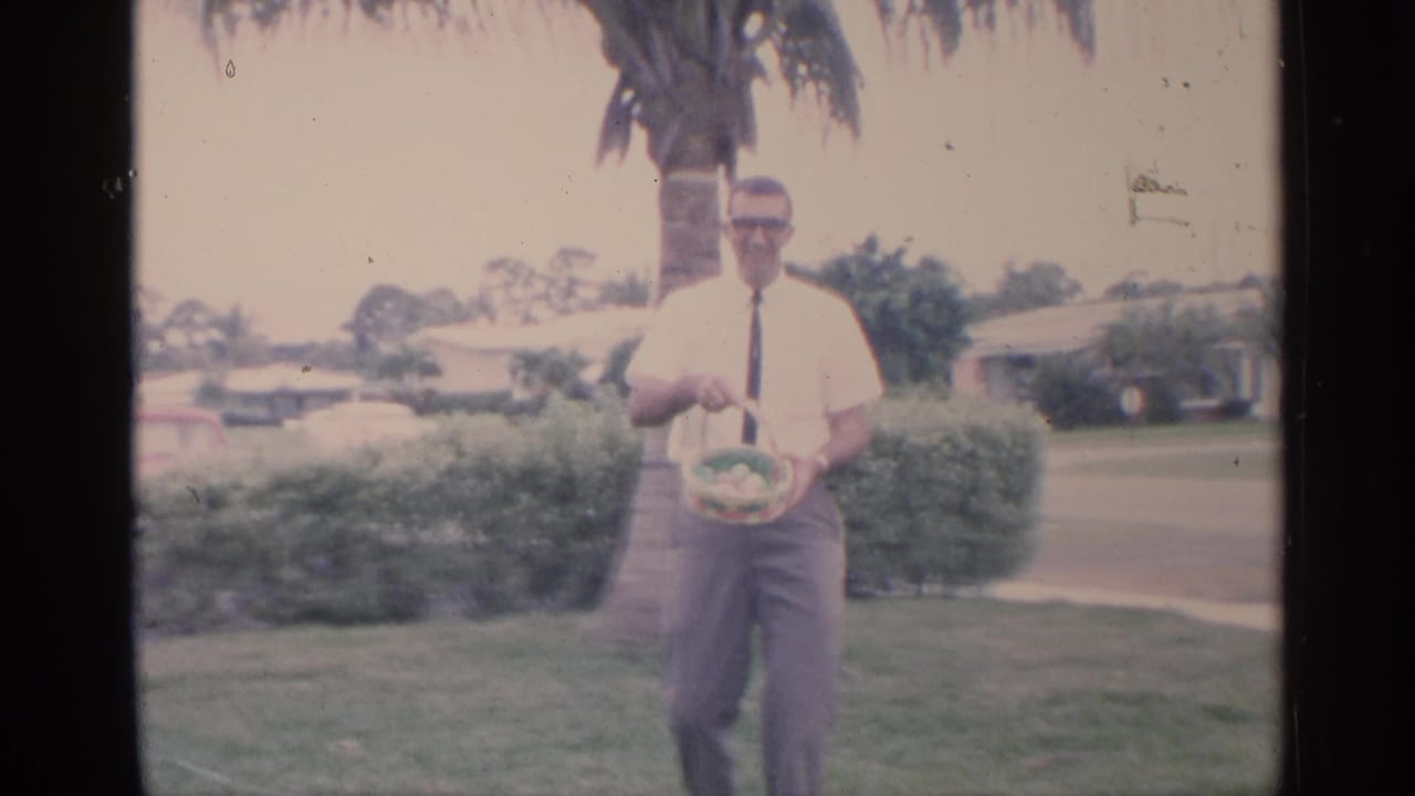 1959 Florida home-movie frame of a man in sunglasses and a dark tie holding an Easter basket on a lawn near palm trees and houses.
