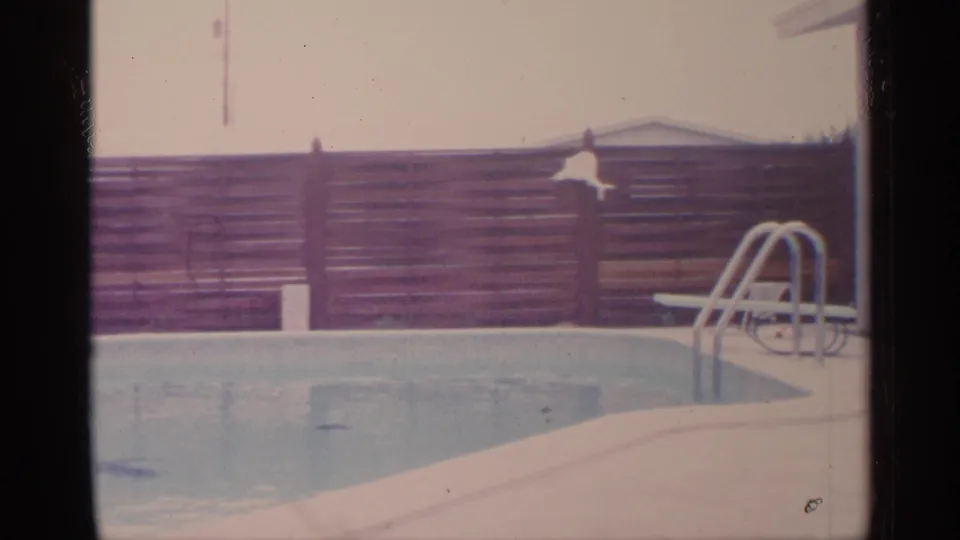1959 Florida home-movie frame of a backyard swimming pool beside a wooden fence, metal ladder, lounge chair, and house exterior.