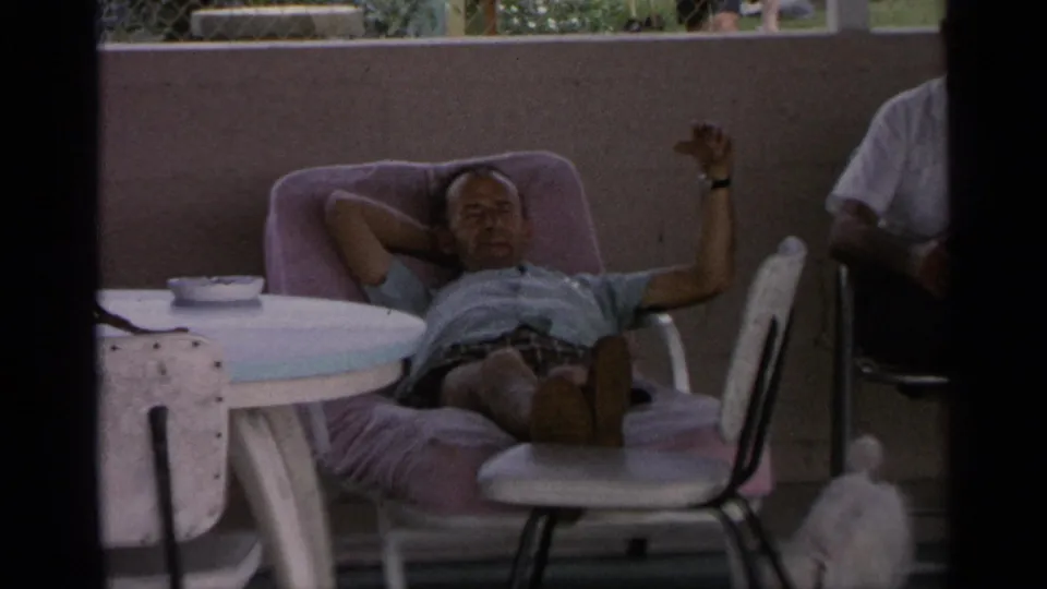 1959 Florida 8mm patio scene with an adult man reclining in a pink lounge chair, a small table, a partial adult figure, and a small white dog.