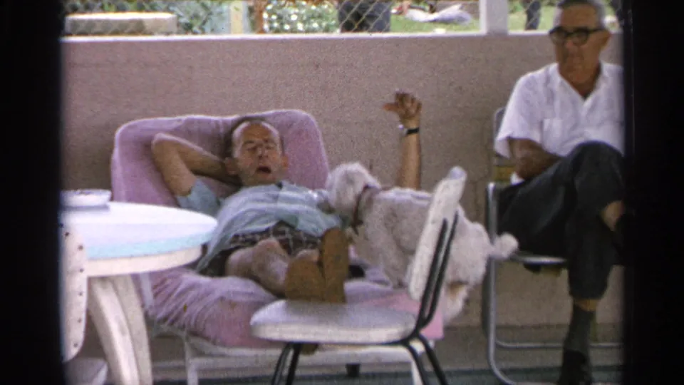 A small white dog reaches the reclining man's lap on a Florida patio while another man sits nearby in the 1959 footage.