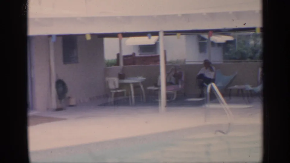 1959 Florida home-movie frame showing an adult seated under a covered patio beside a backyard pool with chairs, tables, and string lights.
