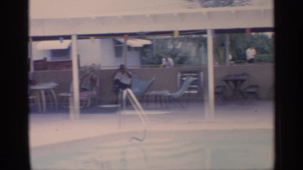 Covered poolside patio with an adult seated near lounge chairs, tables, string lights, and another person in back.