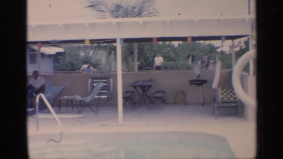 1959 Florida home-movie frame showing patio furniture around a backyard pool, with adults in shade and a pool handrail in the foreground.