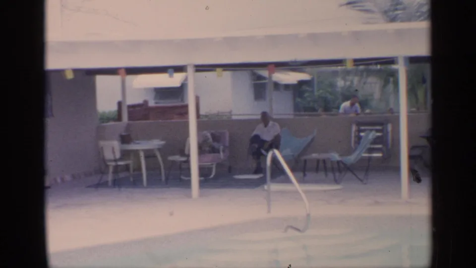 1959 Florida backyard pool frame with an adult seated in the shaded patio area, surrounded by lounge chairs, tables, posts, and string lights.