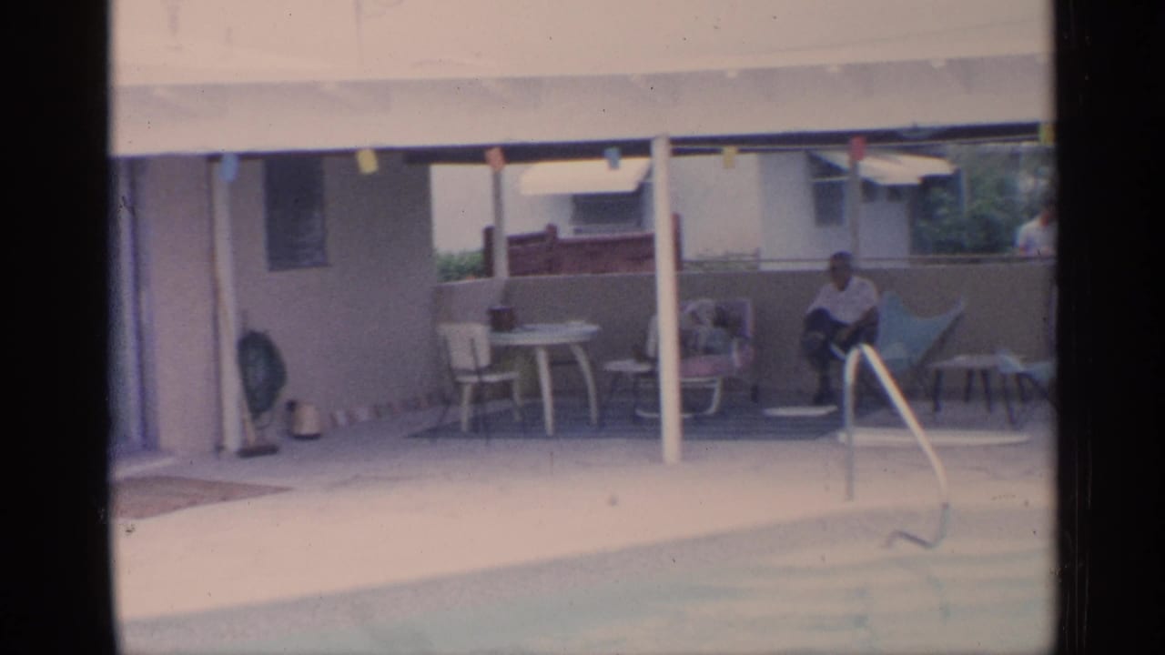 1959 Florida home-movie frame showing a covered patio beside a backyard pool, with an adult seated near chairs and a small table.
