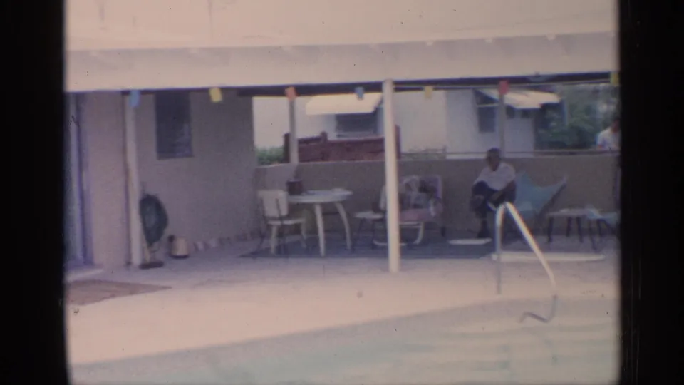 1959 Florida home-movie frame showing a covered patio beside a backyard pool, with an adult seated near chairs and a small table.