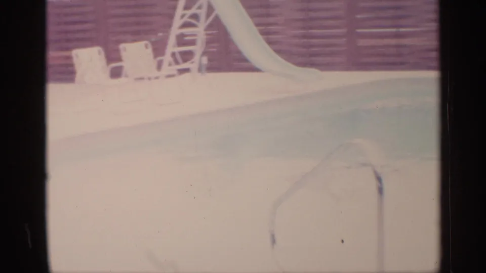 1959 Florida home-movie frame showing a backyard swimming pool with a slide, lounge chairs, brick wall, and handrail.