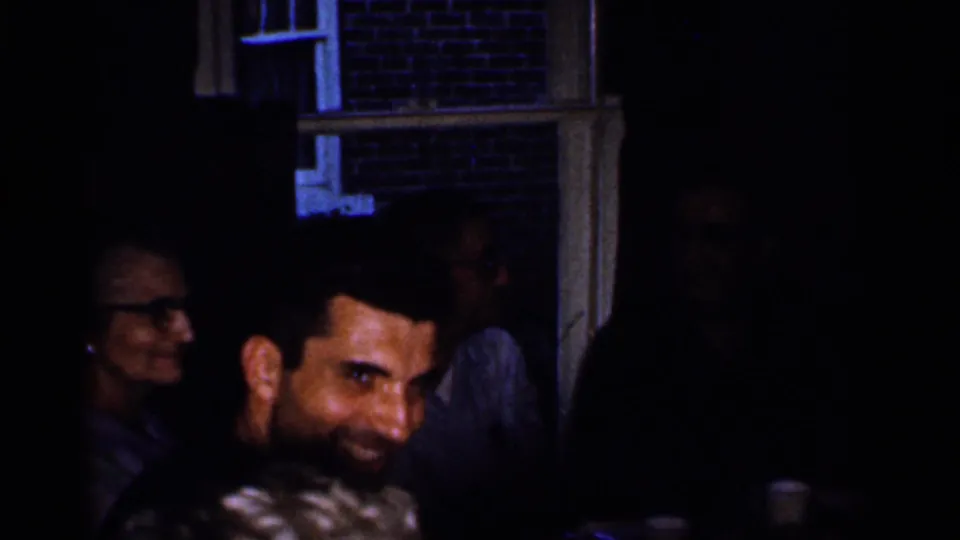 Dimly lit 1959 Florida home-movie party frame with a smiling man in the foreground and guests near a brick exterior.