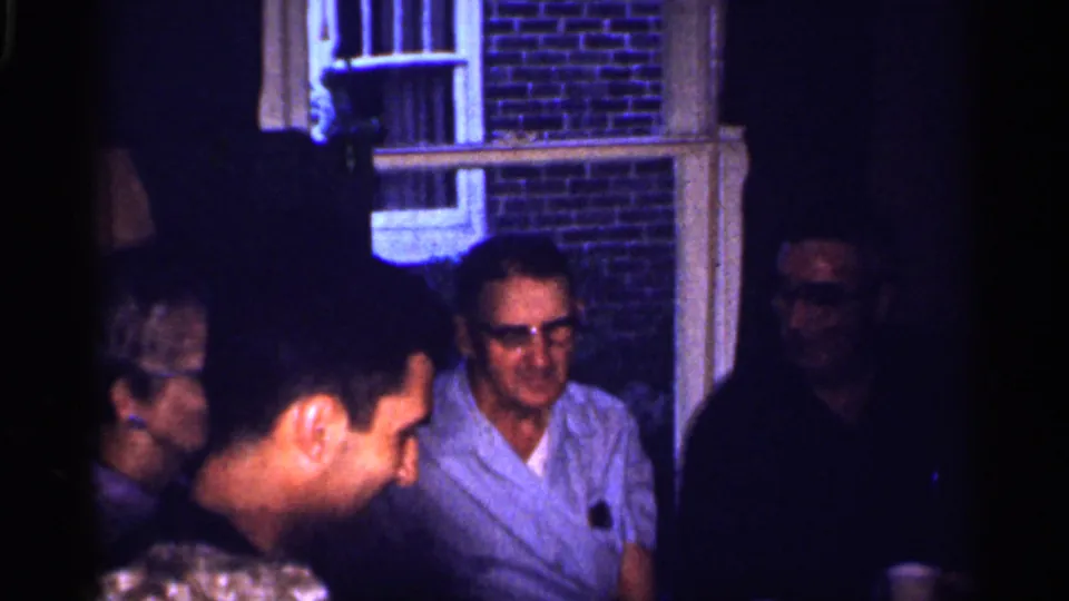 1959 Florida home-movie frame of men conversing at a dim evening party beside a brick house exterior and window.
