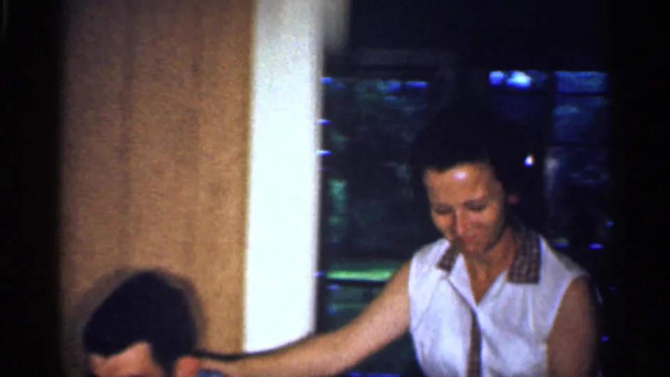 A woman in a white sleeveless blouse with a checkered collar leans toward a seated man inside the Florida home.