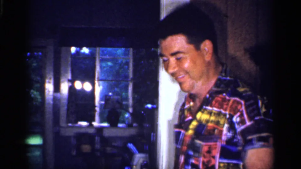 A smiling man in a colorful patterned shirt stands inside the 1959 Florida home with a window and dark garden behind him.