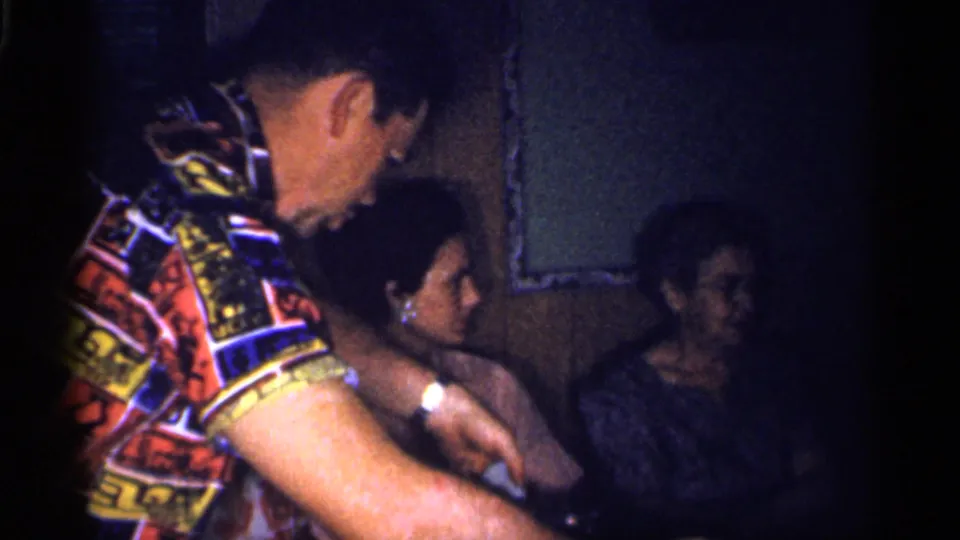 A man in a patterned shirt leans and gestures near seated adults during the indoor Florida home gathering.