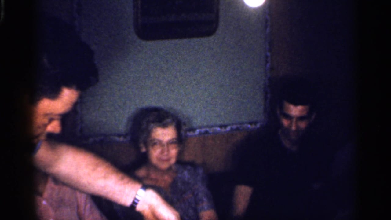 Seated adults in a dim room as a man's arm reaches forward near a woman.