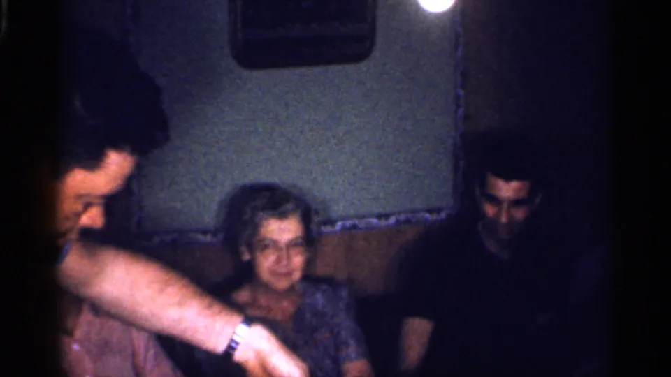 1959 Florida home-movie frame showing adults seated indoors while a man reaches forward near a woman under low light.