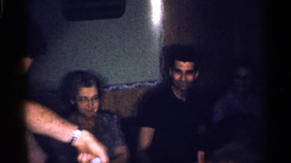 1959 Florida interior frame with a man in a dark shirt, an older woman, and another adult's arm crossing the foreground.