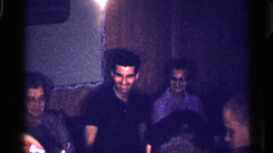 1959 Florida home-movie frame showing seated adults in a dim private room, including a central man in a dark shirt.