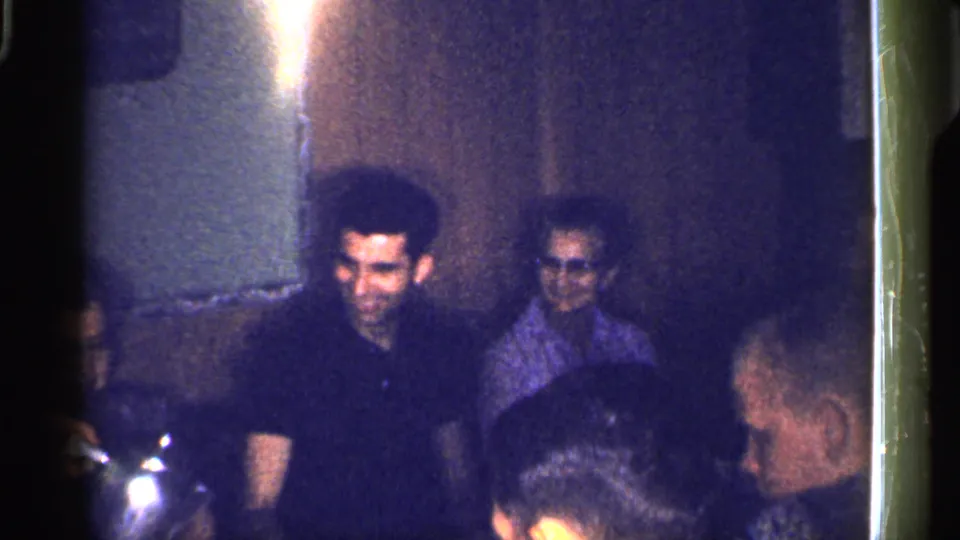 1959 Florida frame showing adults seated in a private interior, with a man in a dark shirt and guests partly obscured in low light.