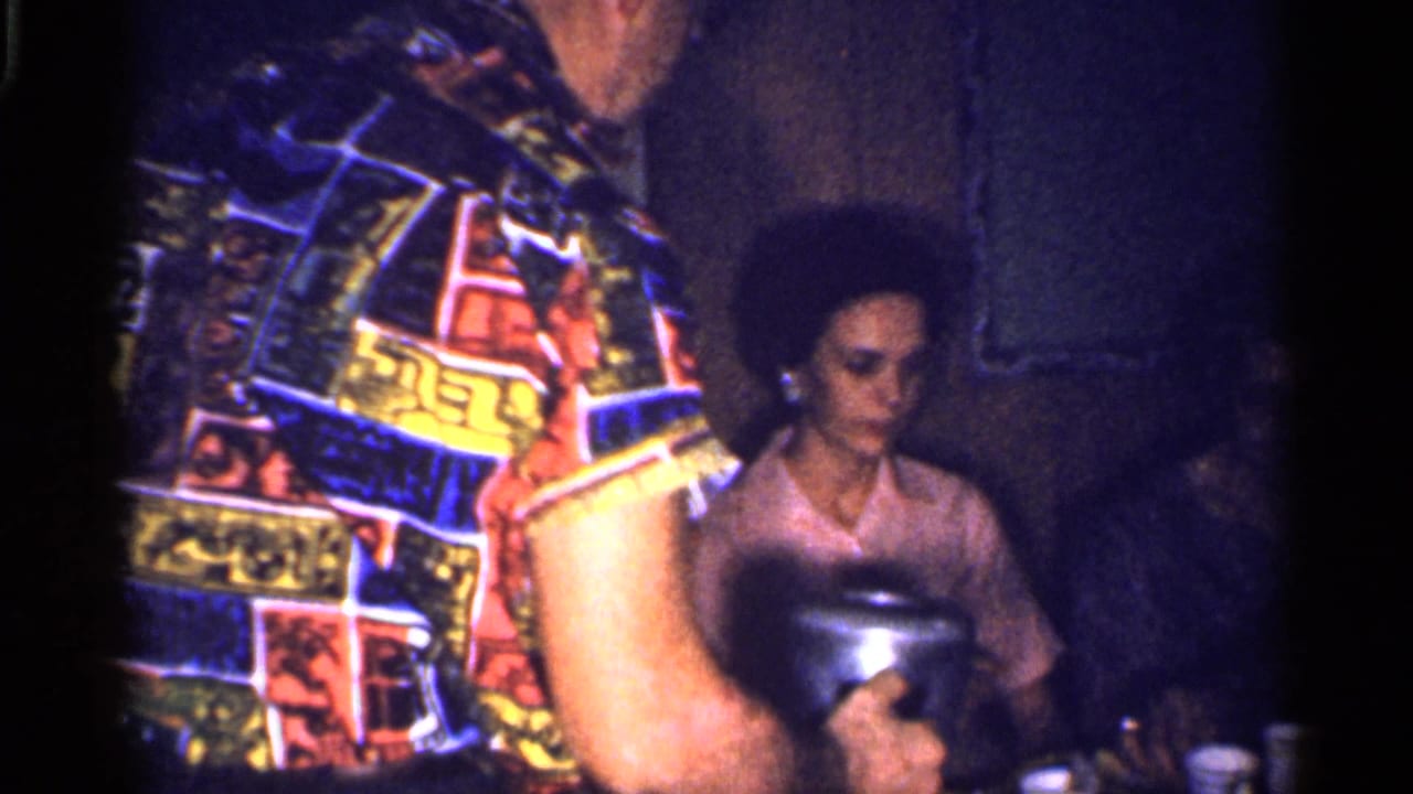 Adults in a dim kitchen, with a man in a patterned shirt foregrounded and a seated woman behind him.
