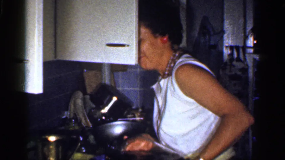 1959 Florida home-movie frame of a woman in a white sleeveless top at a kitchen counter with a metal pitcher and cookware nearby.