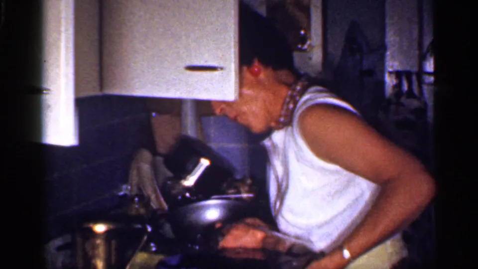 The woman leans toward the counter in a 1959 Florida kitchen, surrounded by metal cookware, cabinets, tile, and household items.