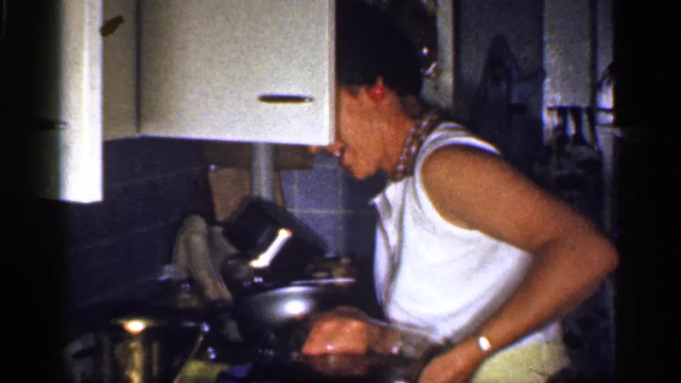 1959 Florida home-movie frame showing the woman at the kitchen counter with a metal pitcher, cookware, wall cabinet, and dark tile.
