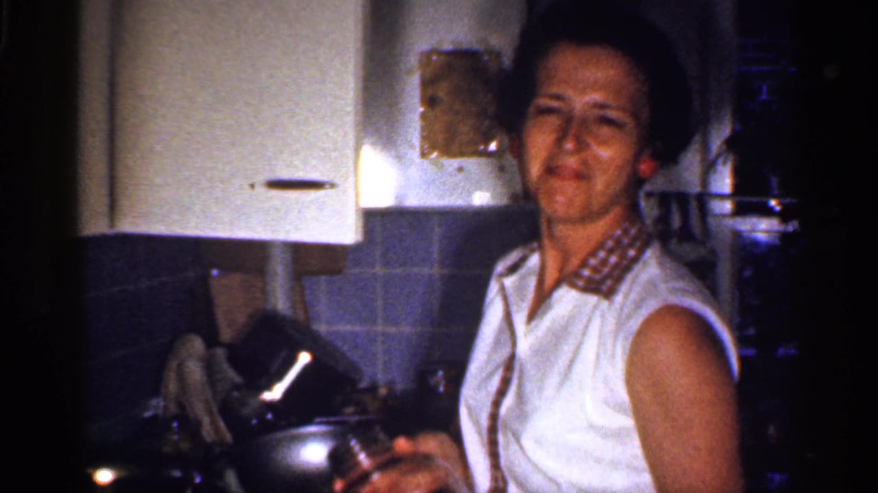 In a 1959 Florida home-movie frame, a woman stands at a kitchen counter near white cabinets, blue tile, and cookware.