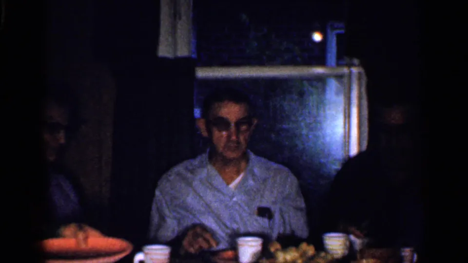 A man with glasses sits at a dining table in a dim Florida home interior, with cups, plates, and relatives partly visible.