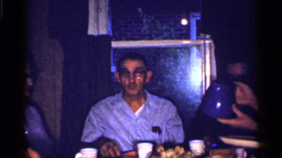 The dining room view shows a seated man with glasses, table cups and food, and a hand offering or holding a pitcher in a private home.