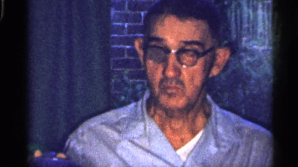 Close 8mm frame of an older man with glasses in a light shirt, seated in a dim domestic dining area in Florida.