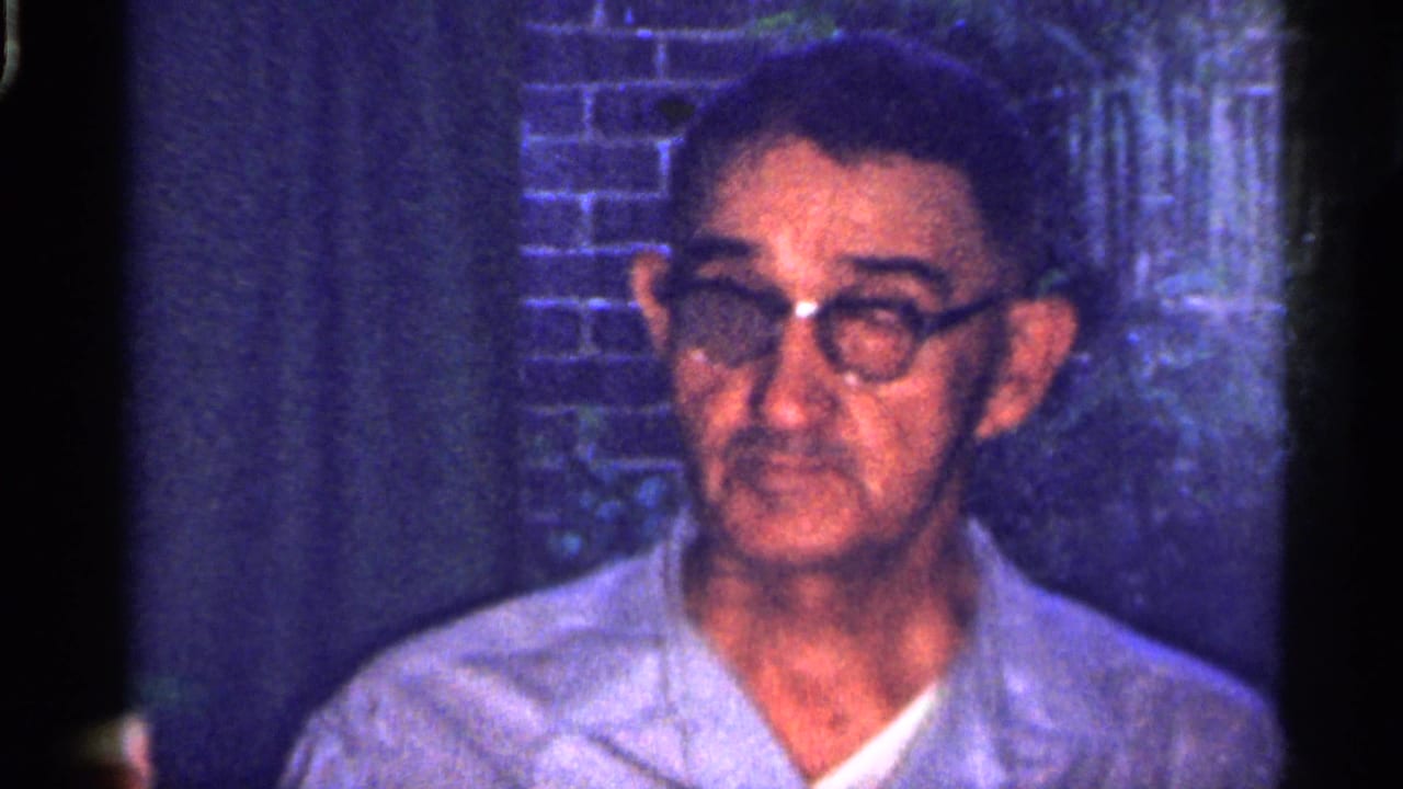 1959 Florida 8mm frame showing an older man in glasses and a light shirt indoors, with brickwork and a dark background behind him.