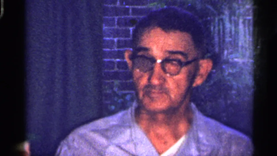 1959 Florida 8mm frame showing an older man in glasses and a light shirt indoors, with brickwork and a dark background behind him.