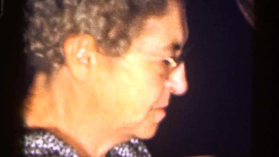1959 Florida 8mm frame showing an older woman in glasses and a pearl necklace in side profile against a dark indoor background.