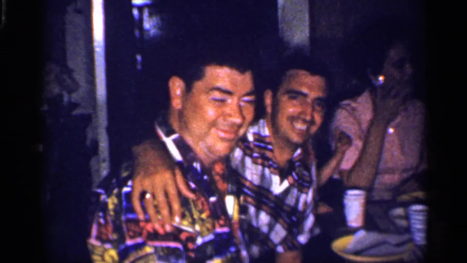 Two men in colorful shirts laugh closely at a dinner table, with another guest visible in the background in 1959 Florida footage.