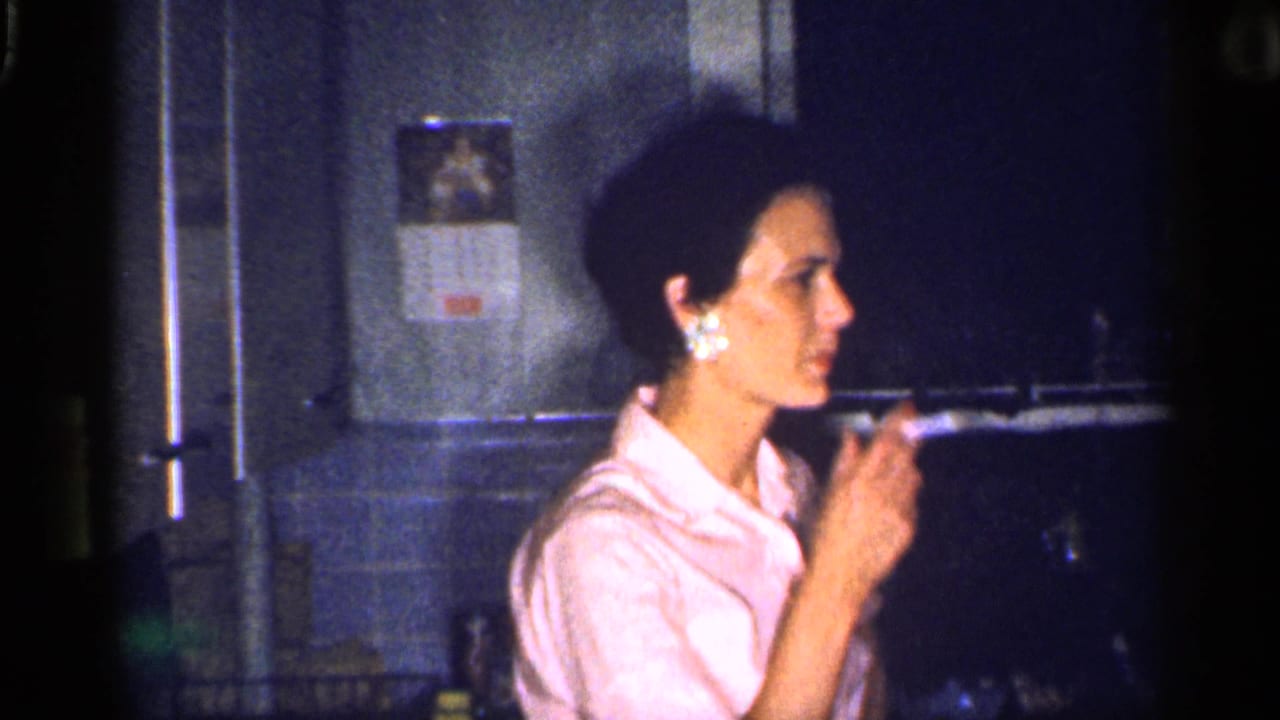 Woman in a pink collared shirt holds a cigarette in a dim kitchen.