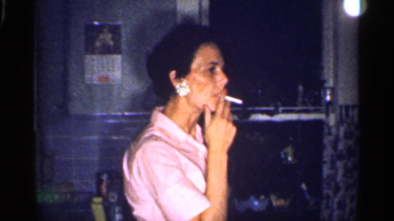 An adult woman in a pink collared shirt smokes in profile near kitchen fixtures, with a calendar and dark counter area behind her in this 1959 Florida home-movie frame.