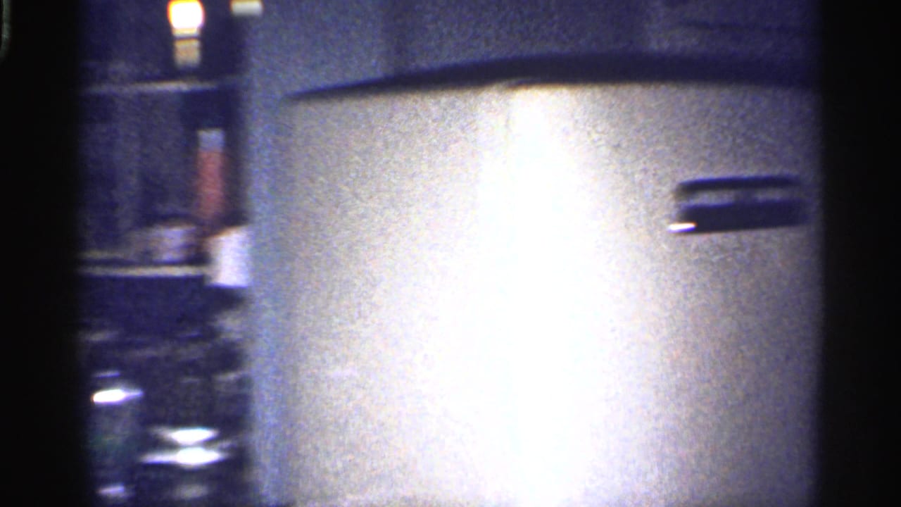 A soft, overexposed kitchen detail shows a pale refrigerator or cabinet surface with nearby shelving and dark film edges visible in the 8mm frame.
